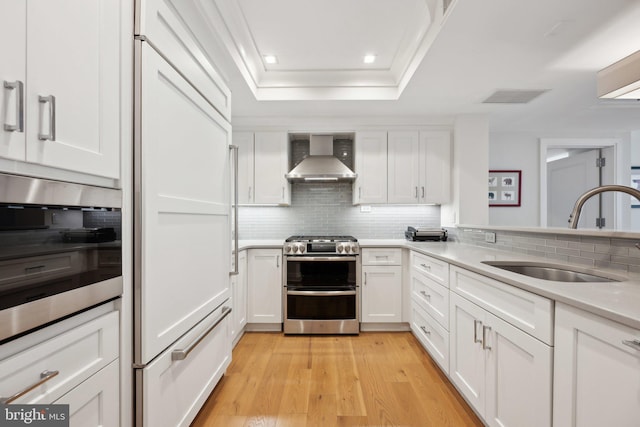 kitchen featuring sink, paneled built in fridge, wall chimney range hood, range with two ovens, and white cabinets