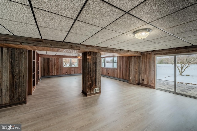 basement with a baseboard radiator, light wood-type flooring, a drop ceiling, and wooden walls