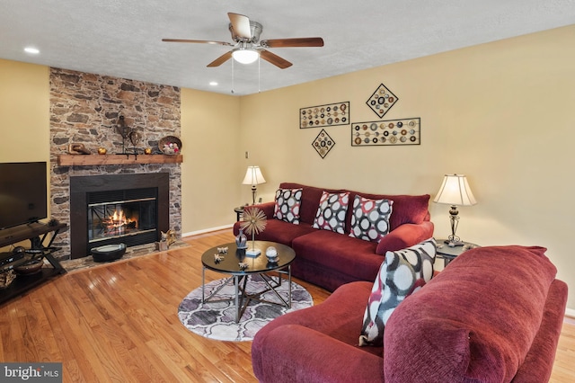 living room with ceiling fan, a stone fireplace, wood-type flooring, and a textured ceiling