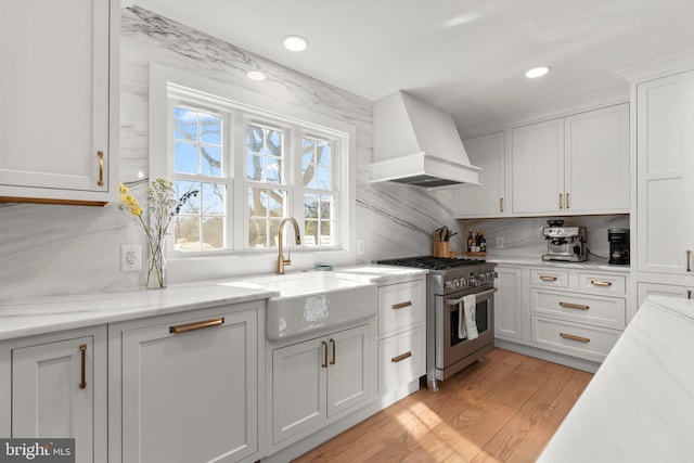 kitchen with premium range hood, sink, white cabinetry, light wood-type flooring, and high end stainless steel range oven