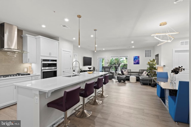 kitchen with sink, white cabinetry, stainless steel appliances, a kitchen island with sink, and wall chimney range hood