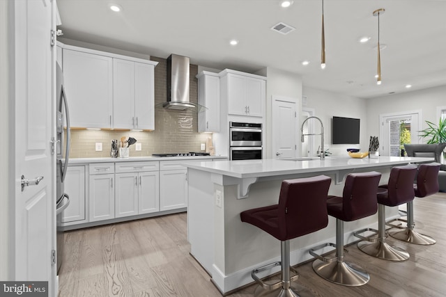 kitchen featuring white cabinetry, wall chimney range hood, stainless steel appliances, and an island with sink