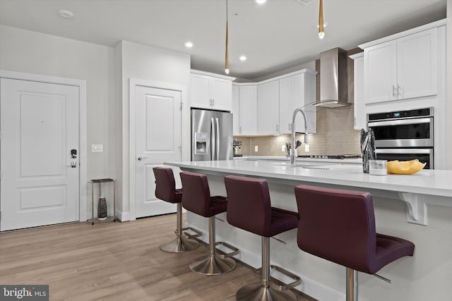 kitchen with wall chimney exhaust hood, a kitchen bar, white cabinetry, light wood-type flooring, and stainless steel appliances