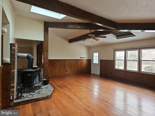 living room with lofted ceiling with skylight, light hardwood / wood-style flooring, a textured ceiling, a wood stove, and ceiling fan