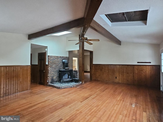 unfurnished living room featuring a textured ceiling, a wood stove, hardwood / wood-style flooring, ceiling fan, and vaulted ceiling with skylight