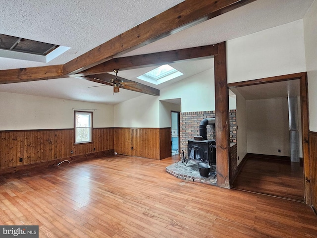 unfurnished living room featuring a textured ceiling, a wood stove, light hardwood / wood-style flooring, ceiling fan, and vaulted ceiling with skylight