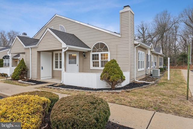 view of front facade with cooling unit, roof with shingles, a chimney, and a front lawn