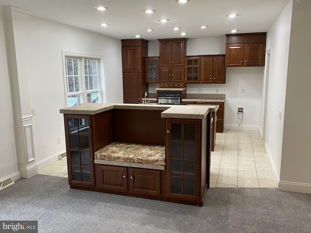 kitchen with dark brown cabinets, tile countertops, an island with sink, and light tile patterned flooring