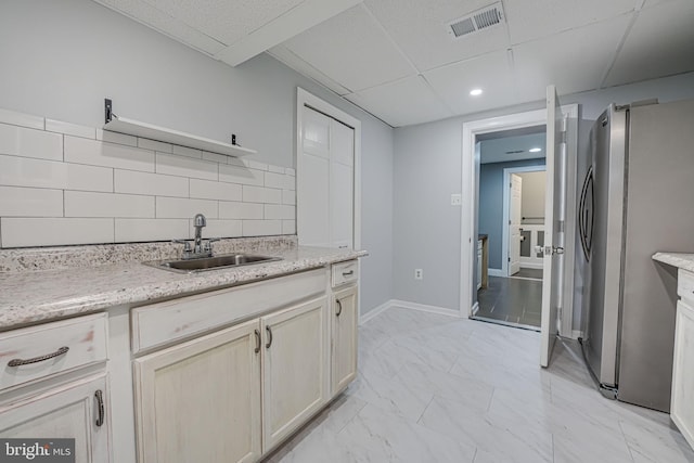 kitchen featuring sink, tasteful backsplash, a paneled ceiling, light stone counters, and stainless steel fridge