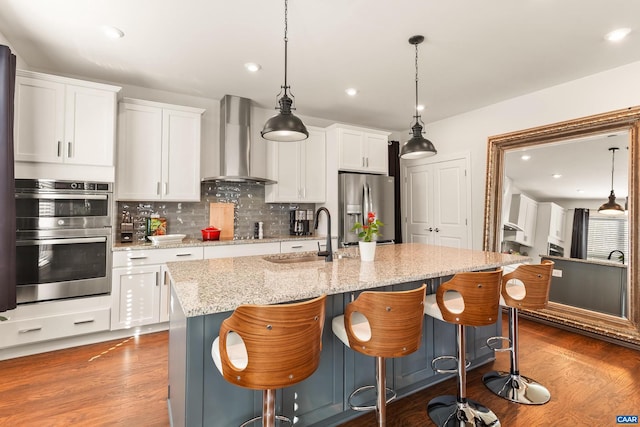 kitchen with white cabinetry, sink, a kitchen island with sink, stainless steel appliances, and wall chimney range hood