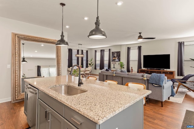 kitchen with sink, a kitchen island with sink, hanging light fixtures, light stone counters, and stainless steel dishwasher