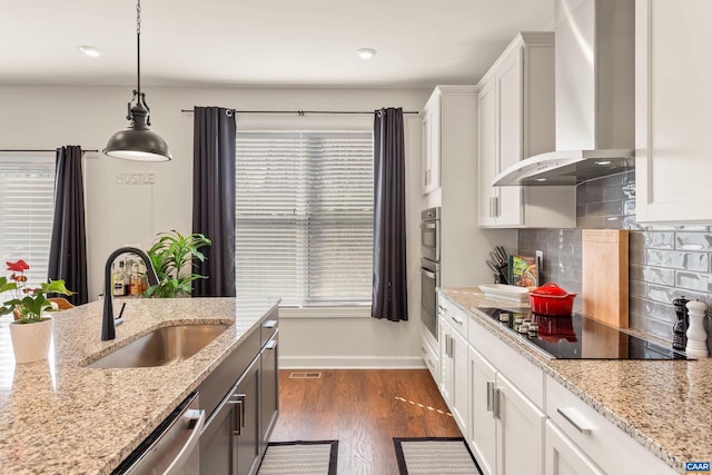 kitchen with wall chimney range hood, sink, hanging light fixtures, stainless steel appliances, and white cabinets