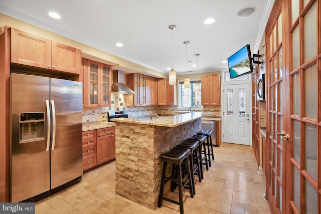 kitchen with pendant lighting, wall chimney range hood, appliances with stainless steel finishes, light stone countertops, and a kitchen island