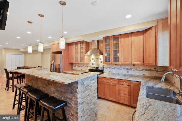 kitchen featuring a kitchen bar, sink, appliances with stainless steel finishes, light stone countertops, and wall chimney range hood