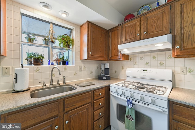 kitchen with under cabinet range hood, a sink, backsplash, brown cabinetry, and white range with gas stovetop