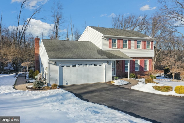 colonial house with a shingled roof, a chimney, a garage, aphalt driveway, and brick siding