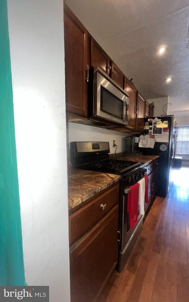 kitchen with appliances with stainless steel finishes, dark brown cabinets, dark hardwood / wood-style floors, a textured ceiling, and dark stone counters