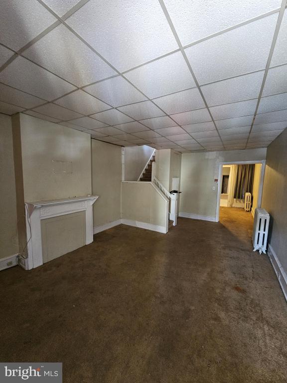 basement with a paneled ceiling, radiator heating unit, and dark colored carpet