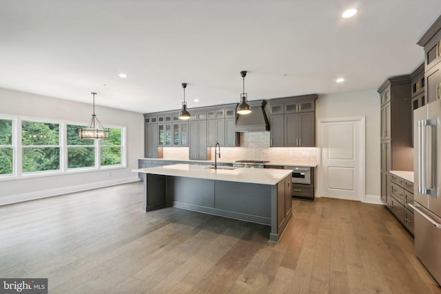 kitchen featuring hardwood / wood-style floors, backsplash, custom exhaust hood, hanging light fixtures, and a kitchen island with sink