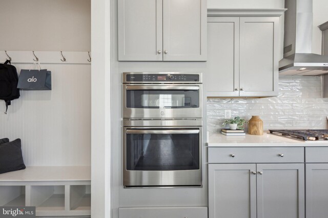 kitchen featuring wall chimney range hood, backsplash, gray cabinets, and stainless steel appliances