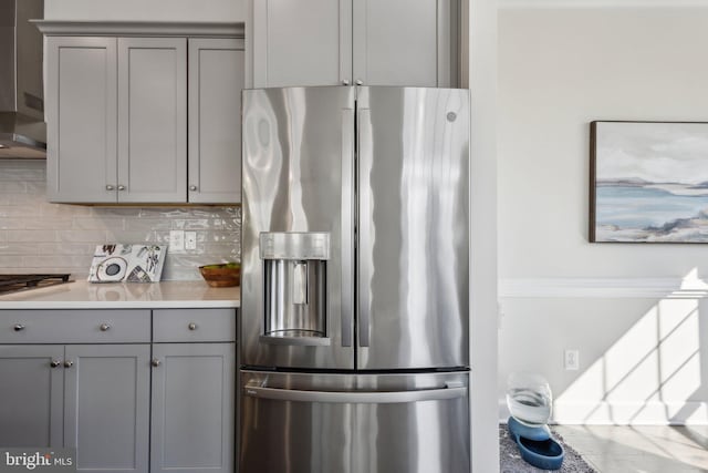 kitchen with stainless steel fridge, gas cooktop, gray cabinetry, backsplash, and wall chimney exhaust hood