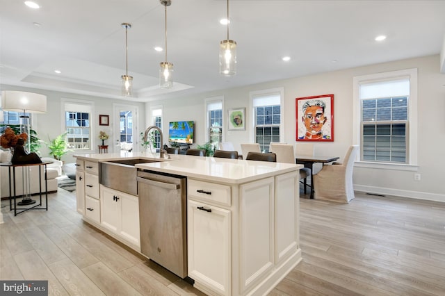 kitchen featuring light wood-style flooring, hanging light fixtures, stainless steel dishwasher, an island with sink, and a raised ceiling