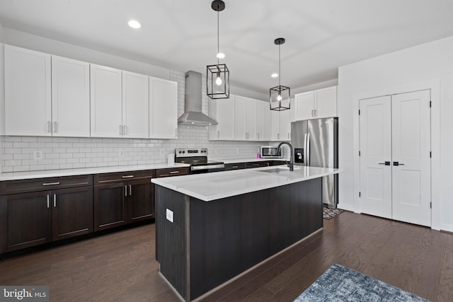 kitchen featuring pendant lighting, an island with sink, stainless steel appliances, dark wood-type flooring, and wall chimney range hood