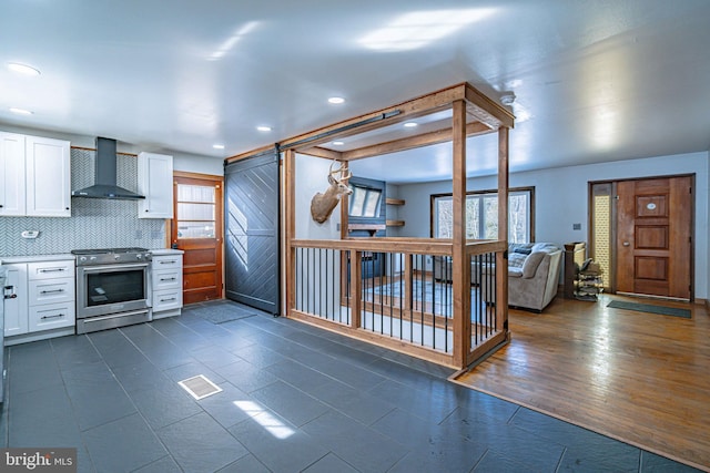 kitchen with wall chimney range hood, stainless steel range, white cabinets, decorative backsplash, and a barn door