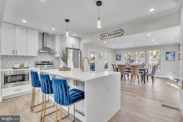 kitchen with stainless steel appliances, light countertops, white cabinetry, an island with sink, and wall chimney exhaust hood