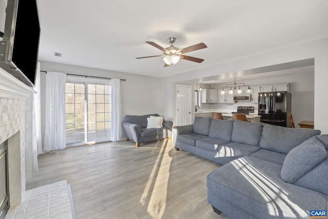 living room with a brick fireplace, ceiling fan, and light wood-type flooring