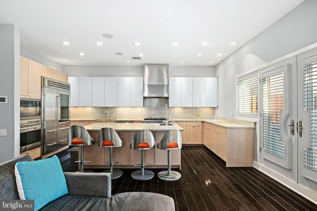 kitchen with a breakfast bar, a kitchen island with sink, built in appliances, decorative backsplash, and wall chimney exhaust hood