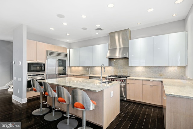 kitchen with exhaust hood, light stone countertops, a kitchen island with sink, and built in appliances