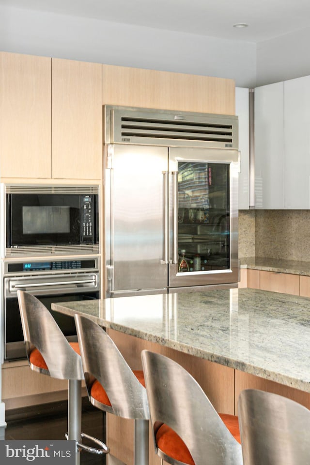 kitchen featuring a breakfast bar, built in appliances, light brown cabinets, light stone countertops, and backsplash