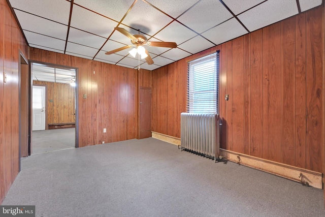 unfurnished room with radiator, a paneled ceiling, ceiling fan, light carpet, and wood walls