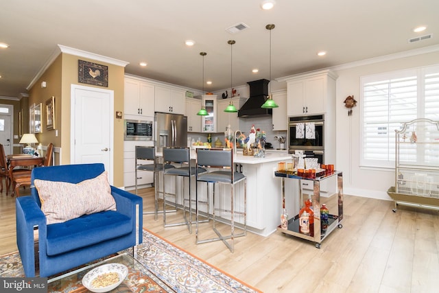 kitchen featuring wall chimney range hood, a breakfast bar area, a kitchen island with sink, hanging light fixtures, and white cabinets