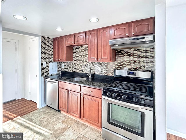 kitchen featuring tasteful backsplash, dark stone countertops, stainless steel appliances, under cabinet range hood, and a sink