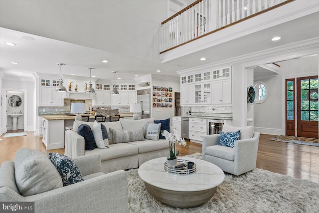 living room with a towering ceiling, beverage cooler, crown molding, wet bar, and light wood-type flooring