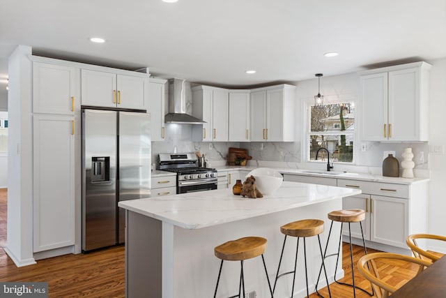 kitchen featuring wall chimney range hood, sink, appliances with stainless steel finishes, hanging light fixtures, and a kitchen island