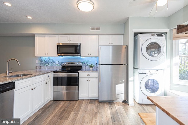 kitchen with stacked washing maching and dryer, appliances with stainless steel finishes, sink, white cabinets, and light hardwood / wood-style flooring