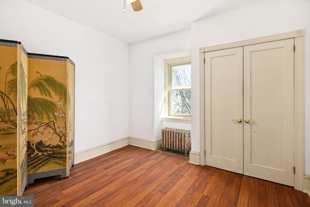 unfurnished bedroom featuring a ceiling fan, dark wood-style flooring, baseboards, and radiator heating unit