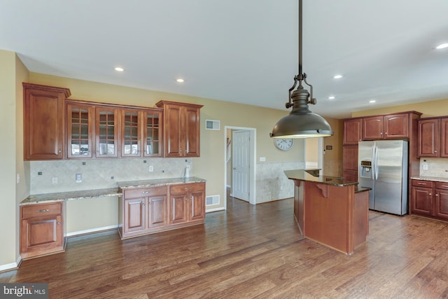 kitchen with light stone counters, hanging light fixtures, stainless steel fridge, dark wood finished floors, and glass insert cabinets