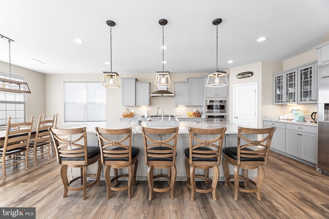 kitchen with decorative light fixtures, gray cabinets, an island with sink, hardwood / wood-style floors, and wall chimney range hood