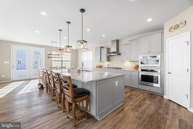 kitchen featuring pendant lighting, wall chimney range hood, gray cabinetry, light stone countertops, and a center island with sink