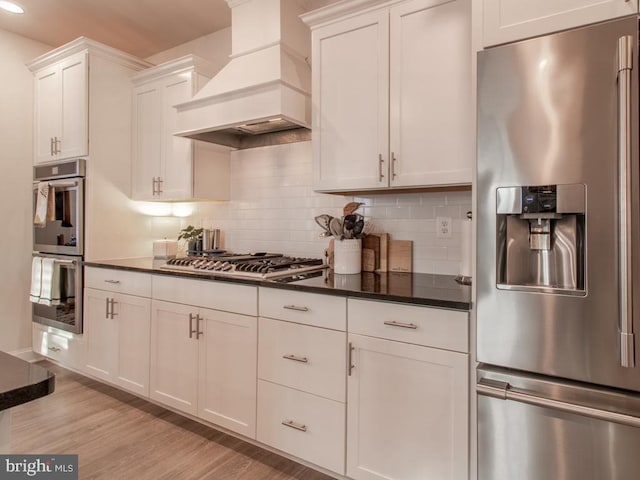 kitchen featuring light hardwood / wood-style flooring, premium range hood, appliances with stainless steel finishes, white cabinets, and decorative backsplash