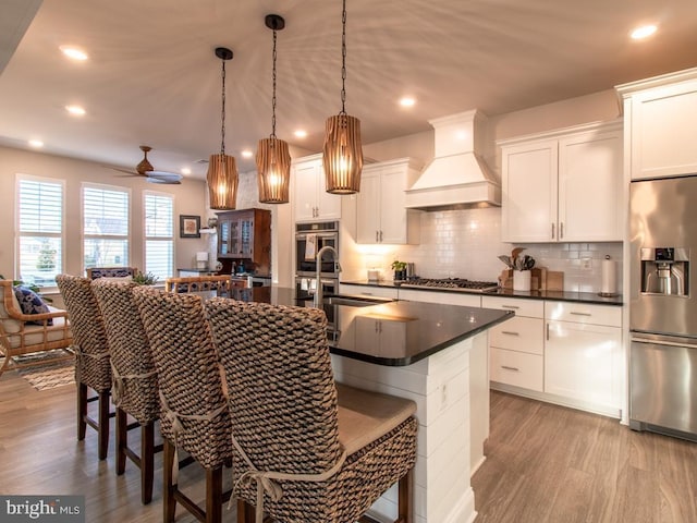 kitchen featuring premium range hood, a kitchen island with sink, hanging light fixtures, white cabinetry, and appliances with stainless steel finishes