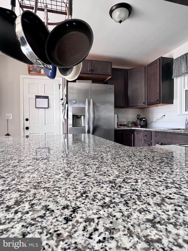 kitchen with dark brown cabinetry, decorative backsplash, light stone countertops, and stainless steel refrigerator with ice dispenser