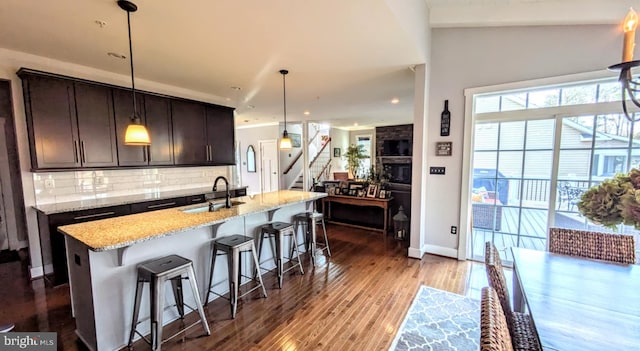 kitchen featuring sink, a kitchen island with sink, dark brown cabinetry, light stone countertops, and decorative light fixtures