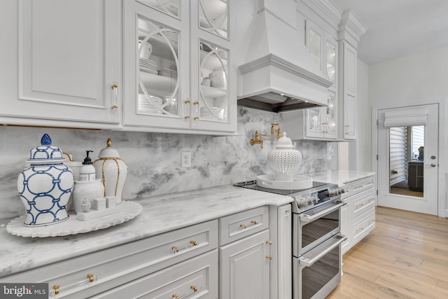 kitchen featuring white cabinets, range with two ovens, custom exhaust hood, light stone counters, and light hardwood / wood-style floors