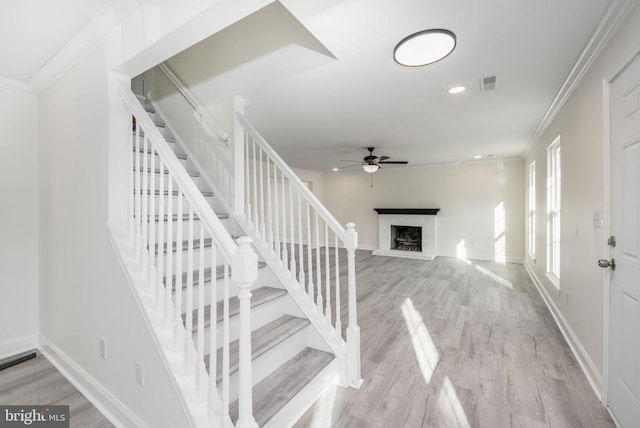 staircase with a brick fireplace, crown molding, wood-type flooring, and ceiling fan