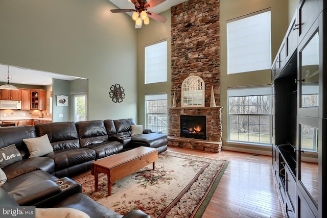 living room with ceiling fan, a stone fireplace, a towering ceiling, and light hardwood / wood-style floors